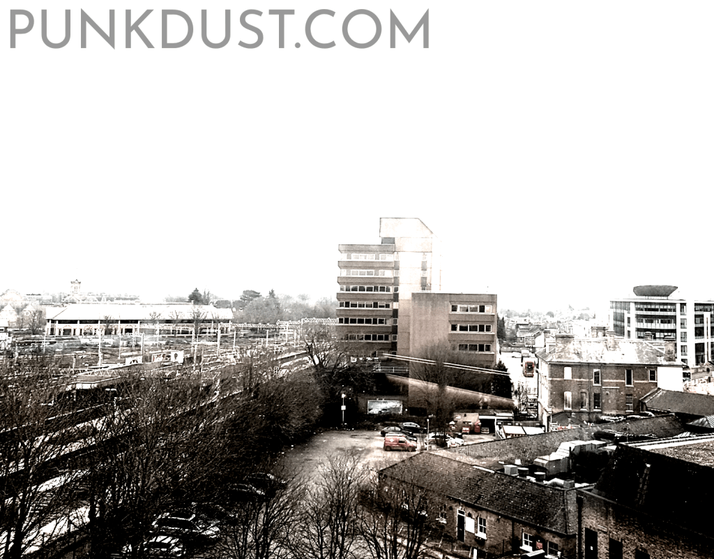Picture of a tired, run-down town centre, with rail lines, car park, and a weird mix of 20th century architecture.